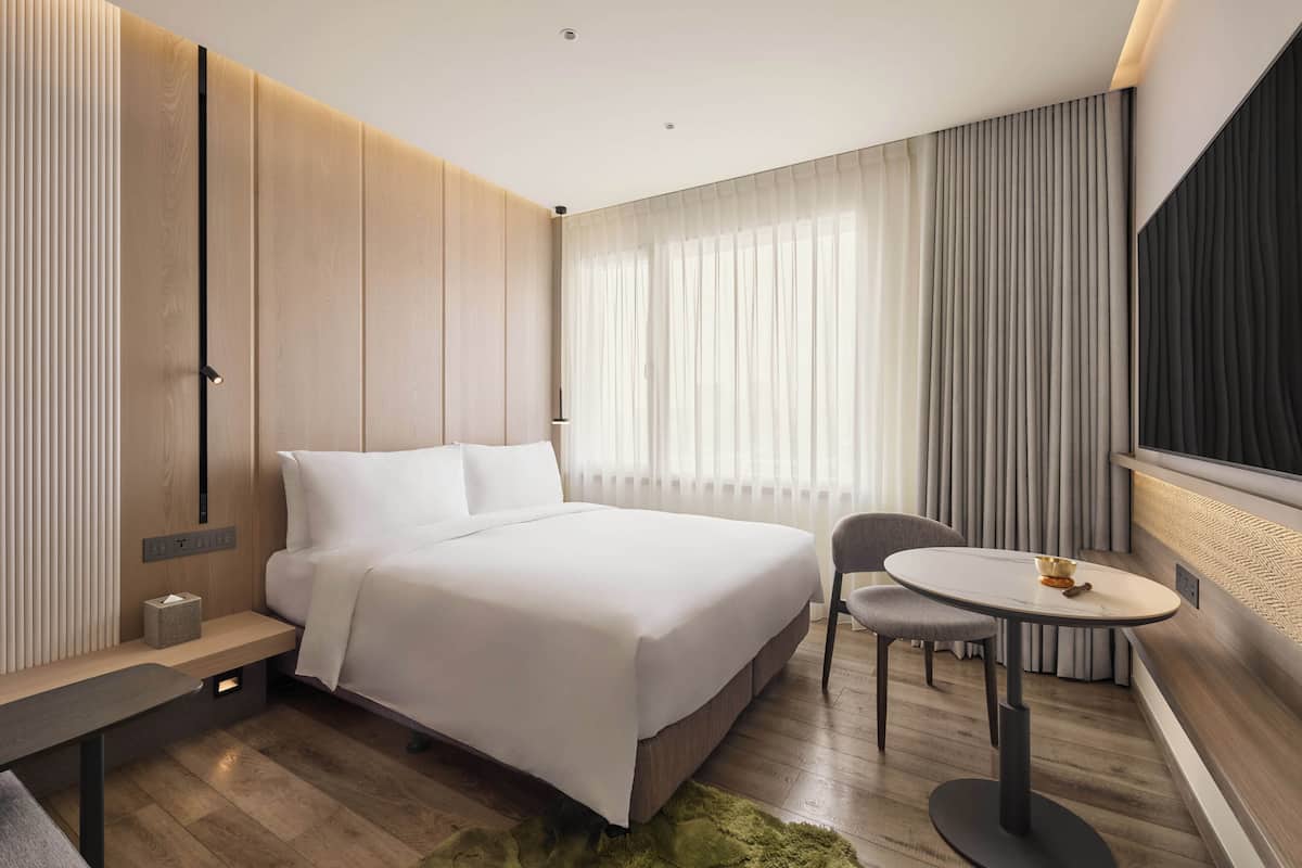 A modern hotel room with a large bed, bedside table, chair, and a small round table.