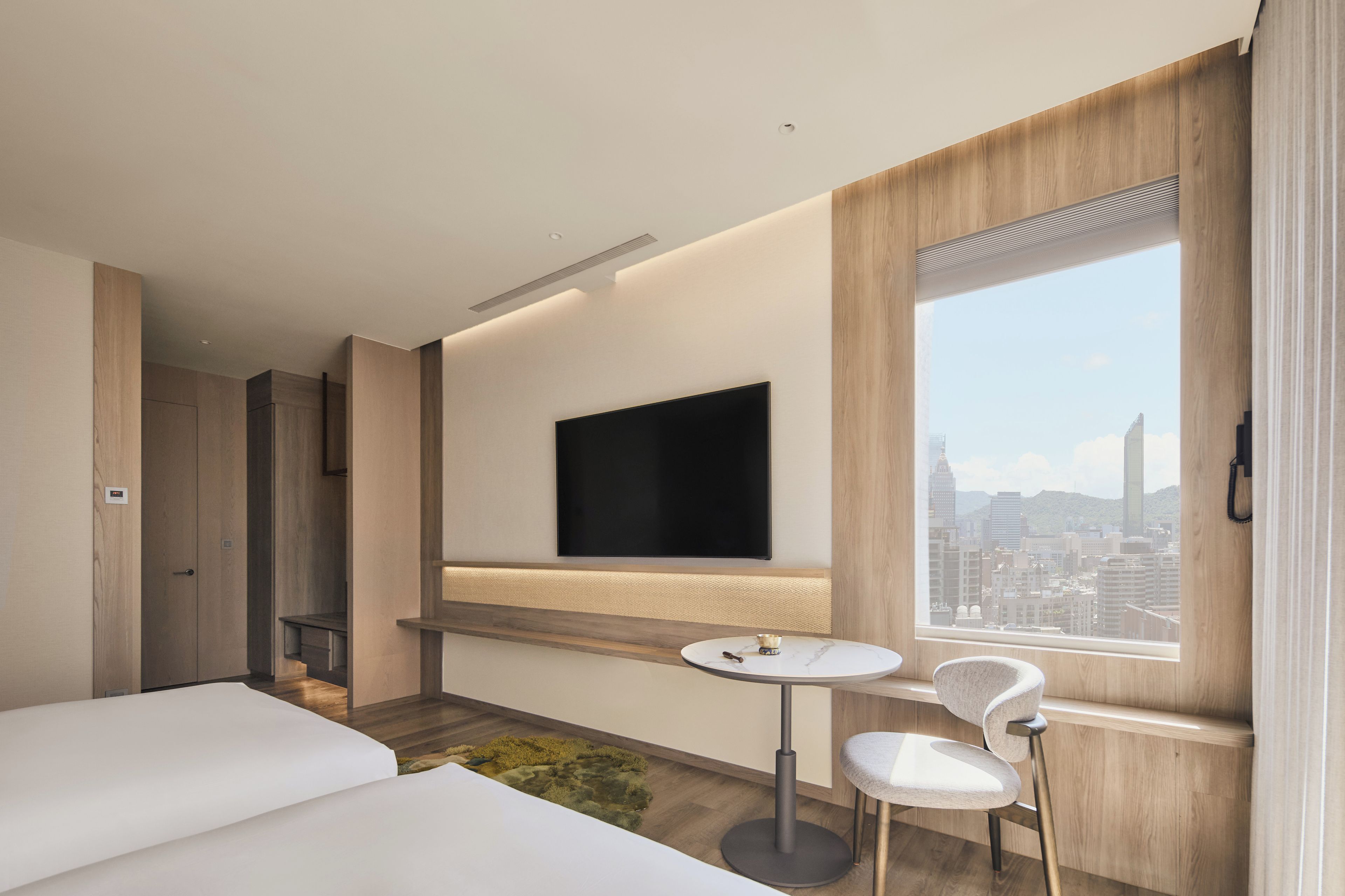 A modern hotel room with a bed, a small table with a chair, a flat-screen TV mounted on the wall, and a large window offering a city view.