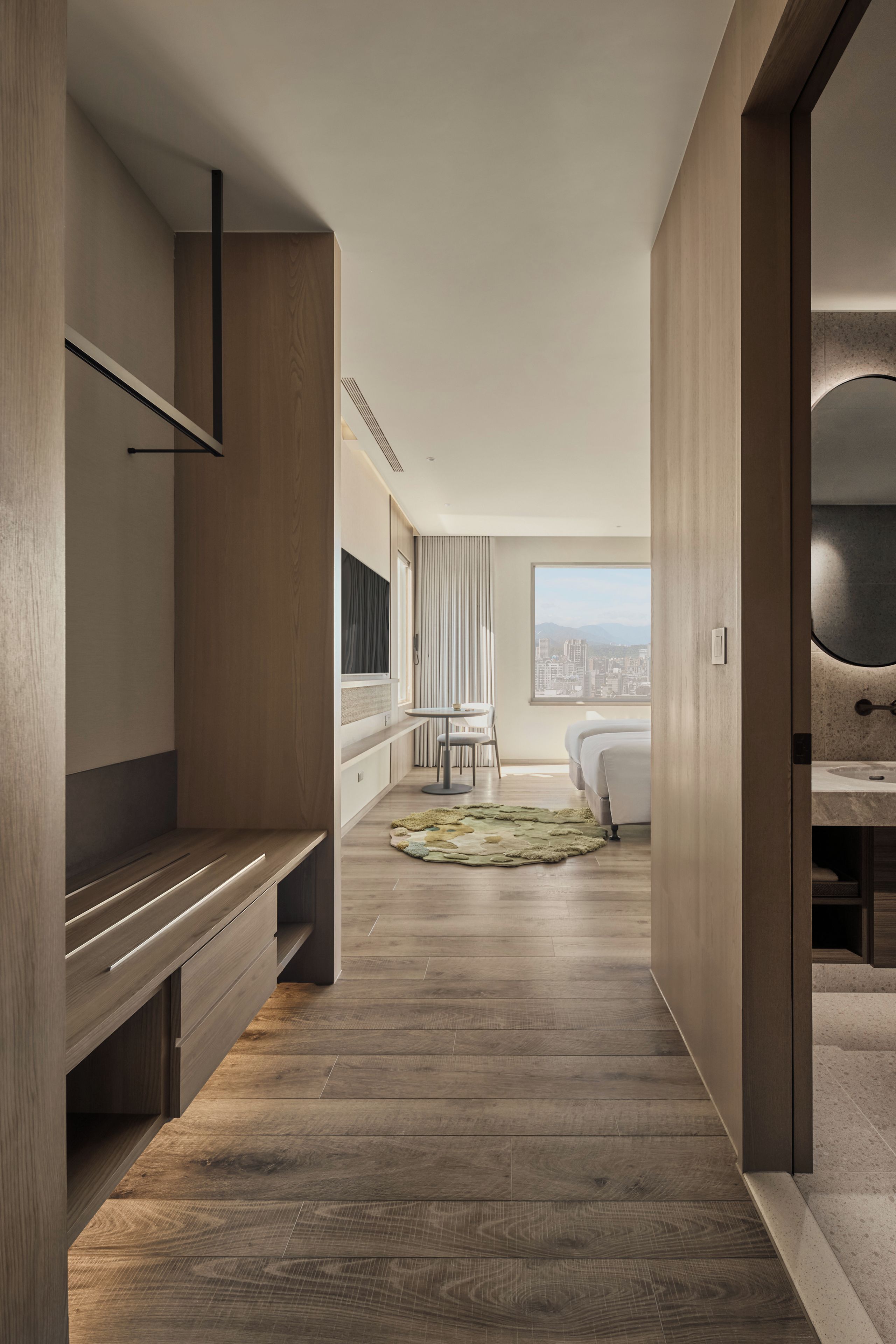 A modern hotel room with a wooden floor, a bed, a desk, and a view of the cityscape.