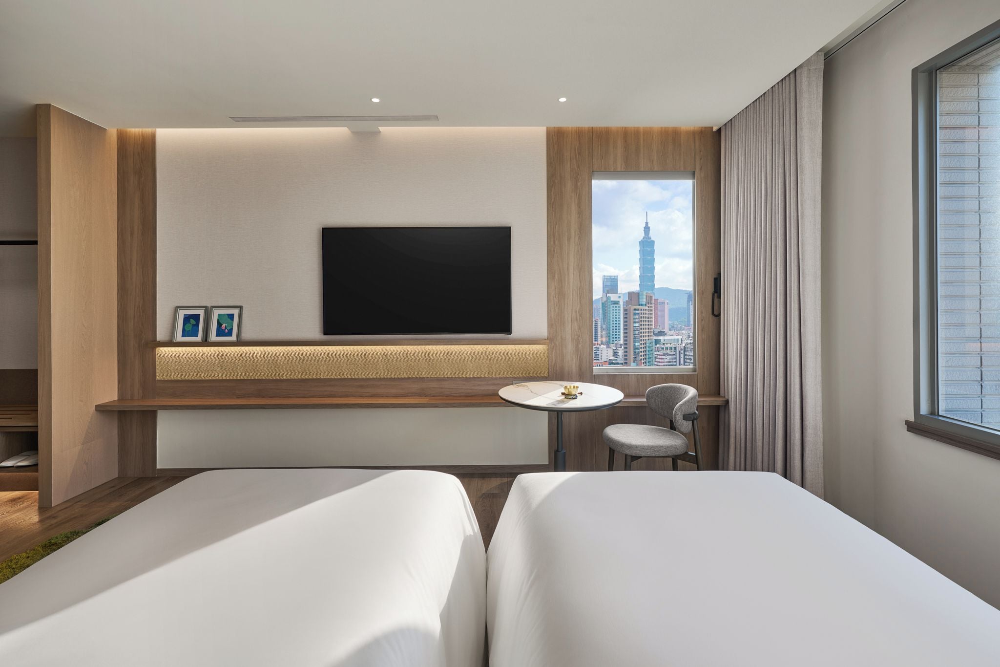 A modern hotel room with two beds, a flat-screen TV mounted on the wall, a small round table, and a city view through the window.