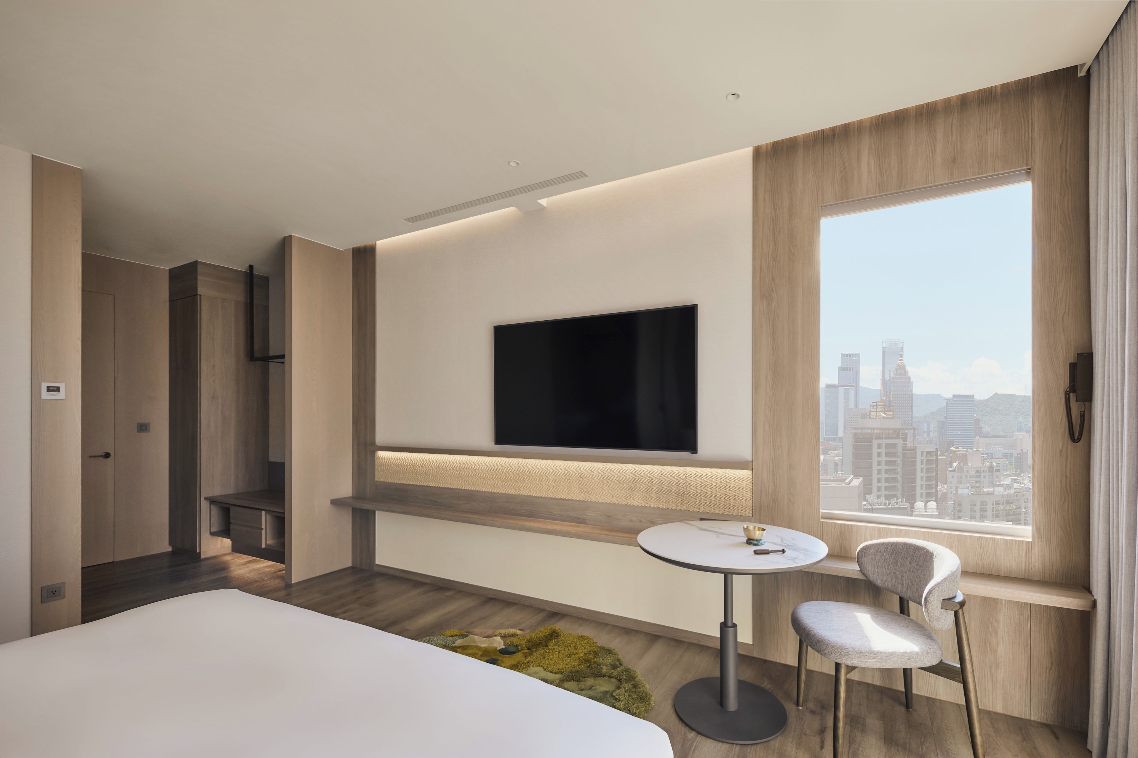 Una habitación de hotel moderna con una cama, un televisor fijado a la pared, una pequeña mesa redonda con una silla y vistas a la ciudad a través de un gran ventanal.