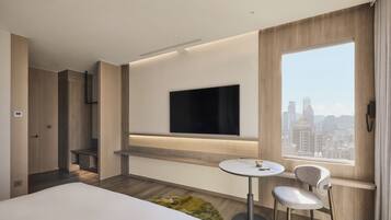 Una habitación de hotel moderna con una cama, un televisor fijado a la pared, una pequeña mesa redonda con una silla y vistas a la ciudad a través de un gran ventanal.