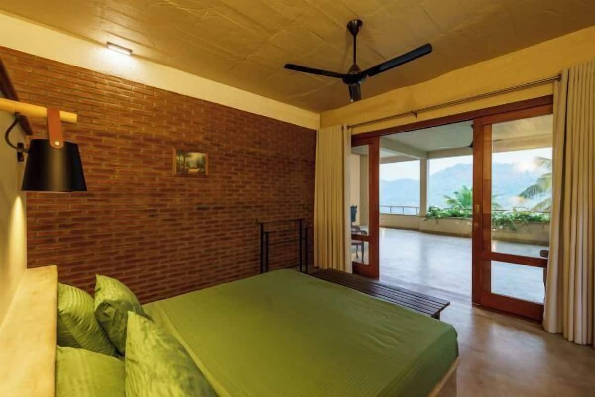 Une pièce avec un lit, un ventilateur de plafond, un mur en briques et une vue sur l’extérieur, visible à travers une porte vitrée.