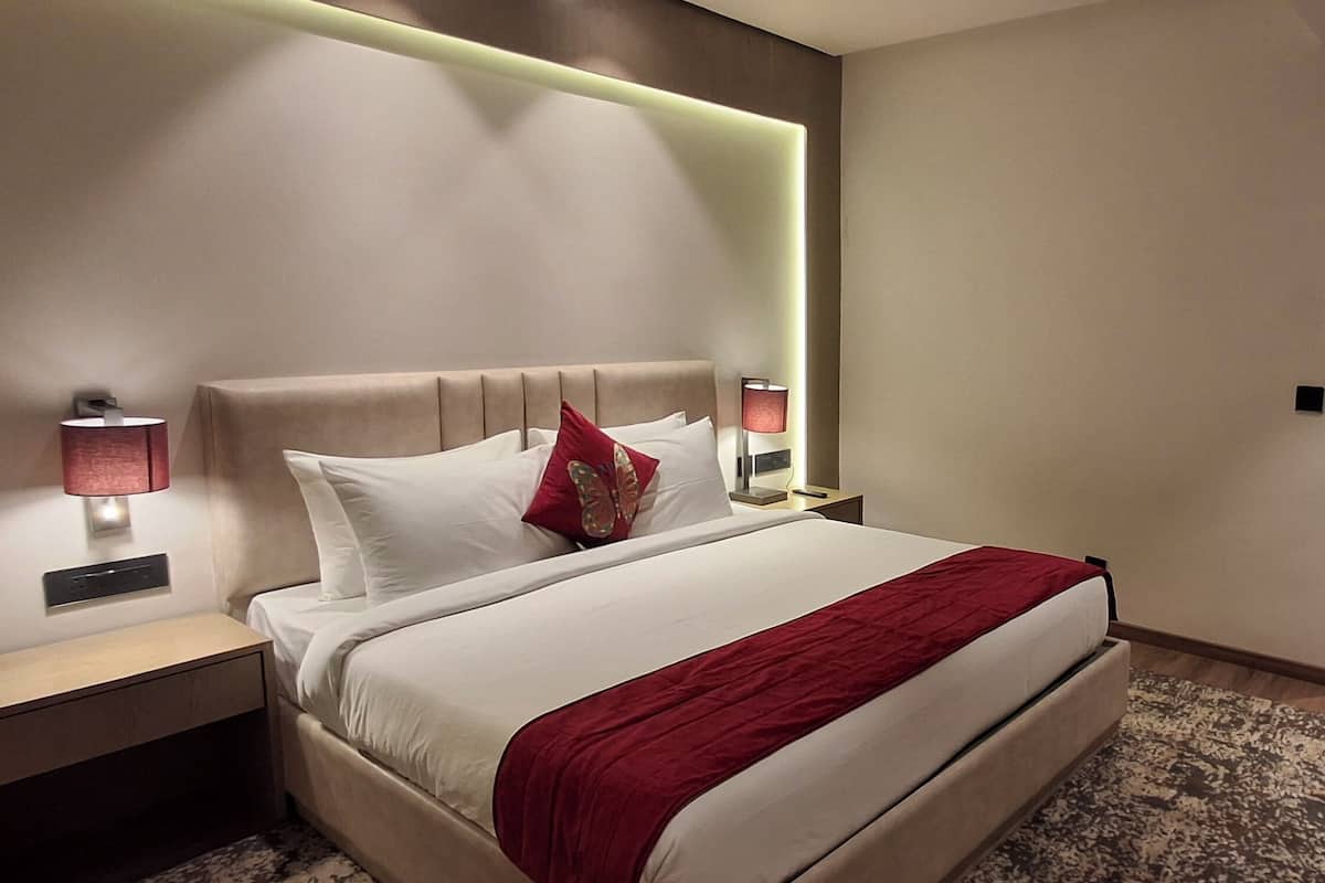 Ein modernes Hotelzimmer mit einem großen Bett, zwei Nachttischlampen, einer roten Tagesdecke und Blick durch Vorhänge aus dem Fenster.
