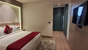 A hotel room with a large bed, a flat-screen TV mounted on the wall, a bedside table with a lamp, and a view of a hallway with curtains. - Rosastays Hansi (Hansi)