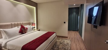 Habitación de hotel con una cama grande, un televisor de pantalla plana montado en la pared, una mesita de noche con lámpara y vistas a un pasillo con cortinas.