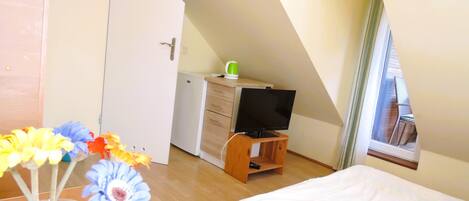 A bedroom with a bed, a TV on a wooden stand, a small desk with a chair, and a window with a view.