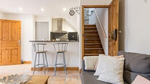 Una sala de estar moderna con un sofá, dos taburetes altos, una mesa de centro de madera y una escalera que lleva a una zona de cocina.
