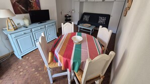 A dining area with a striped tablecloth, white chairs, a television on a cabinet, and a painting on the wall. - Collioure T2 Apartment with Terrace and Parking (Collioure)