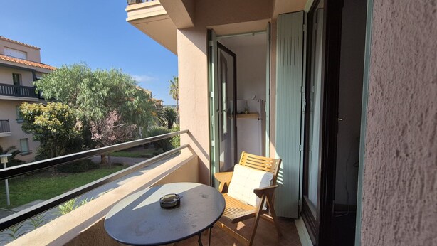 A balcony with a small table and a chair, a sliding door, and a view of the surrounding buildings and greenery. - Collioure T2 Apartment with Terrace and Parking (Collioure)