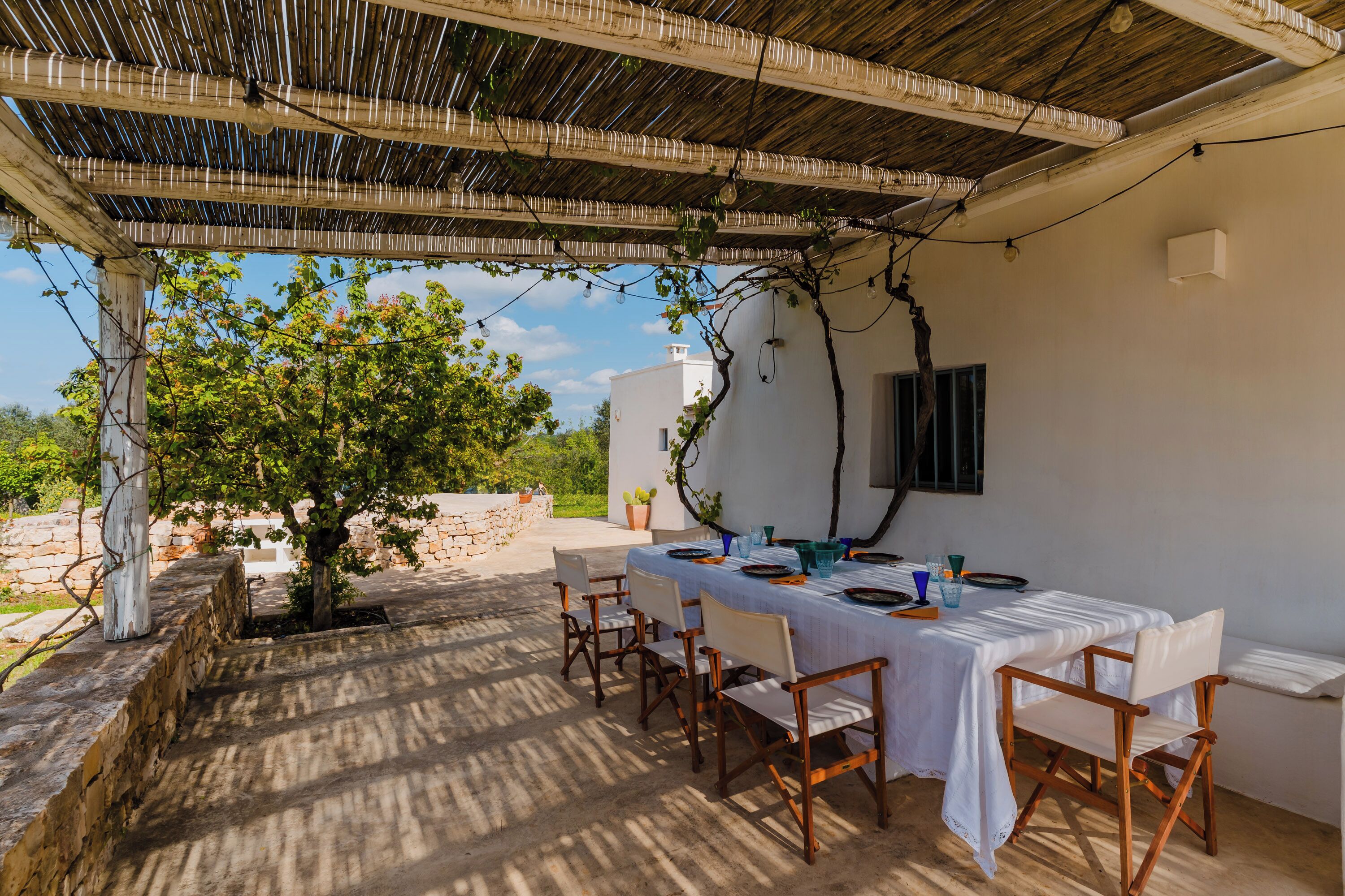 Uma área de refeições ao ar livre com pergolado, uma mesa posta para quatro pessoas e vista para uma árvore e um edifício ao fundo.