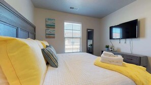 Ein Schlafzimmer mit Bett, Flachbildfernseher, Fenster mit Jalousien, Nachttisch mit Lampe und Wandfernseher.