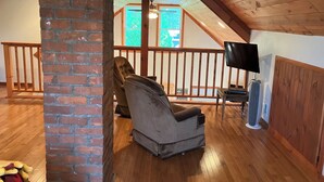 Une pièce mansardée chaleureuse avec un plafond en bois, un mur en briques, une rambarde en bois, un fauteuil inclinable, un téléviseur à écran plat et un ventilateur sur pied.