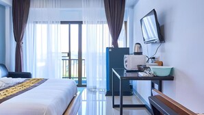 A hotel room with a bed, a desk with a computer and a kettle, a TV on the wall, and a view of the outdoors through large windows. - Belvilla Chiangmai near Lanna Hospital (Chiang Mai)