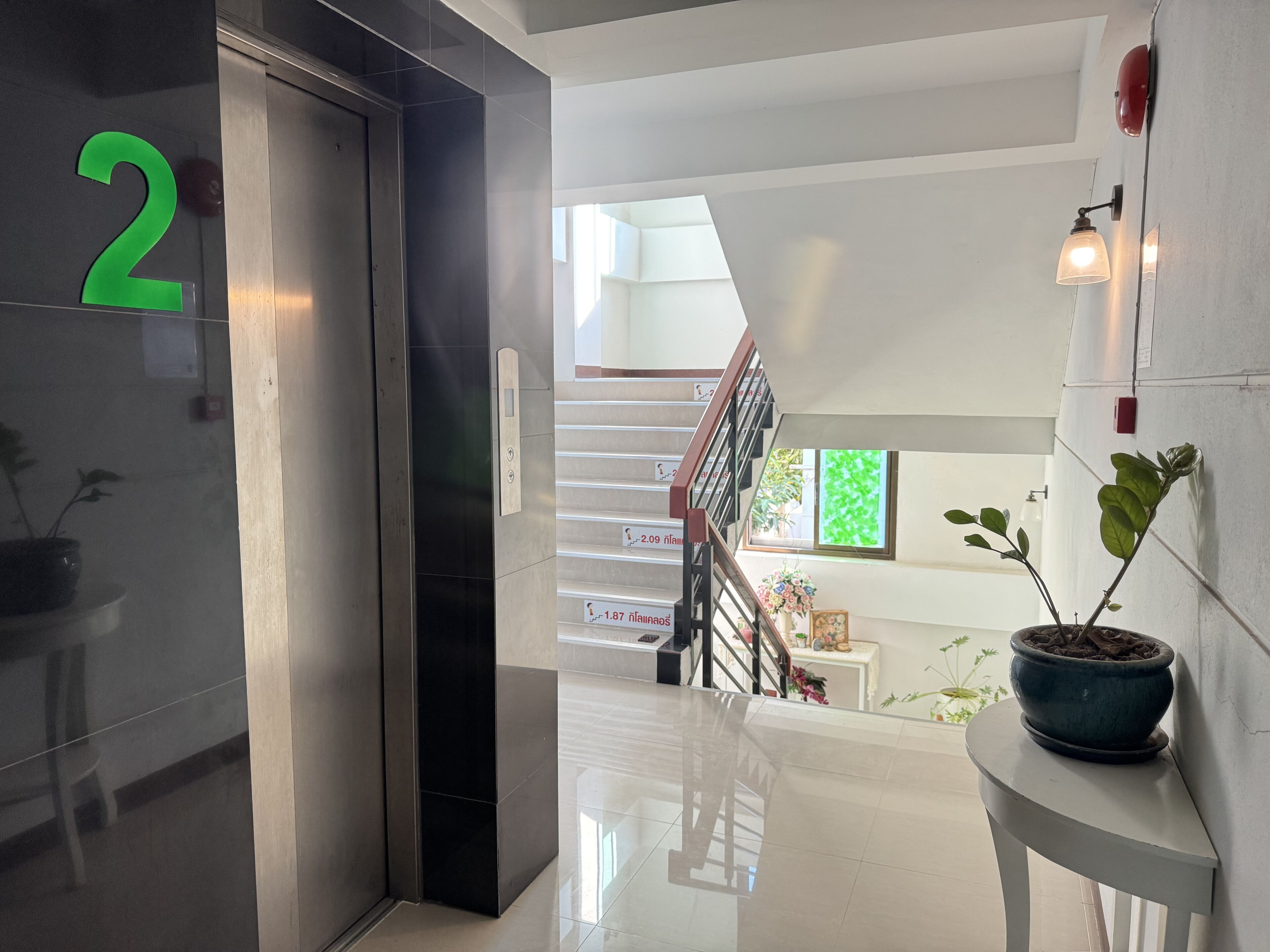 A modern interior with a metallic elevator marked with the number 2, a staircase with a railing, and a potted plant on a white table.