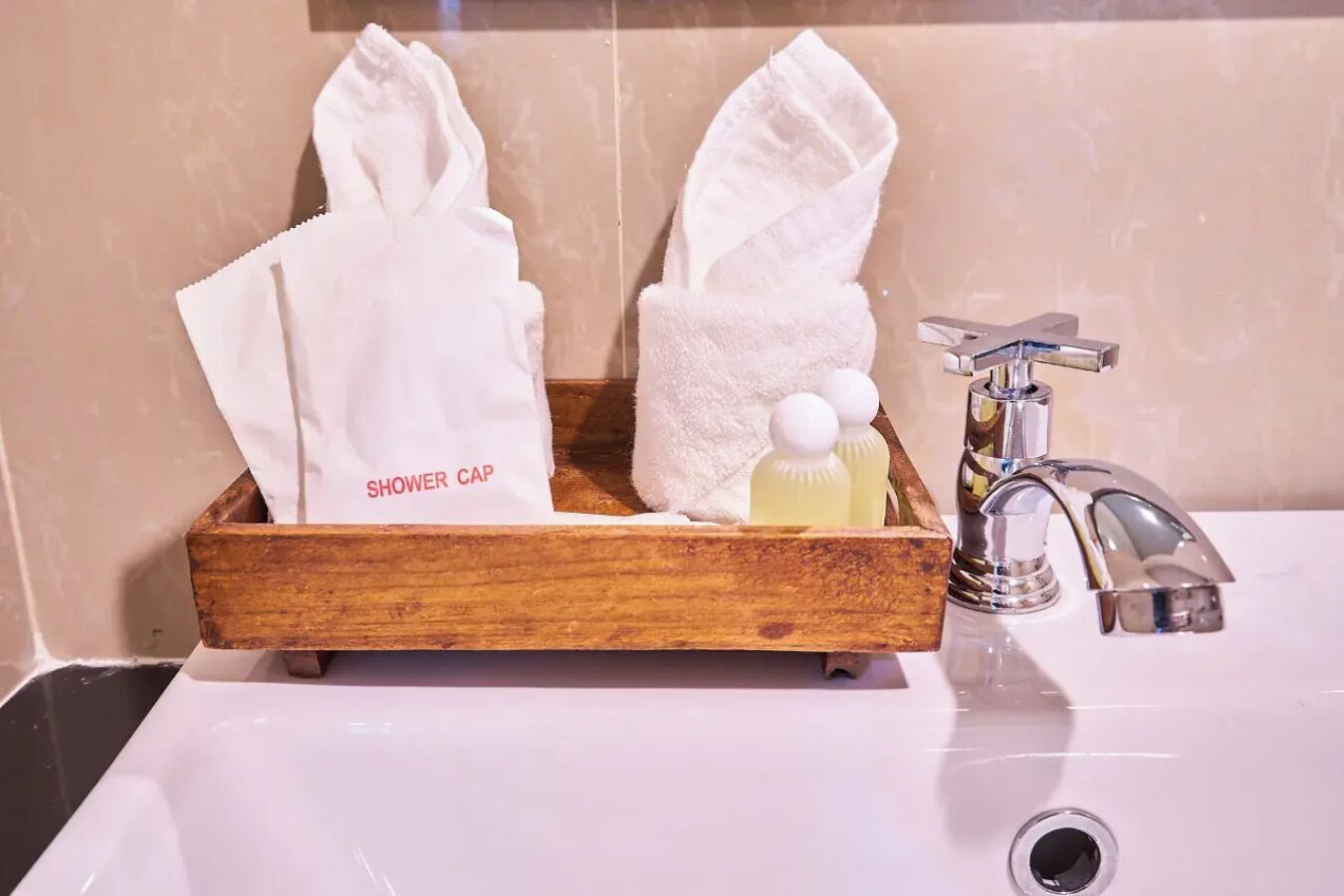A wooden tray with a shower cap, towels, and a bottle of soap on a bathroom sink.