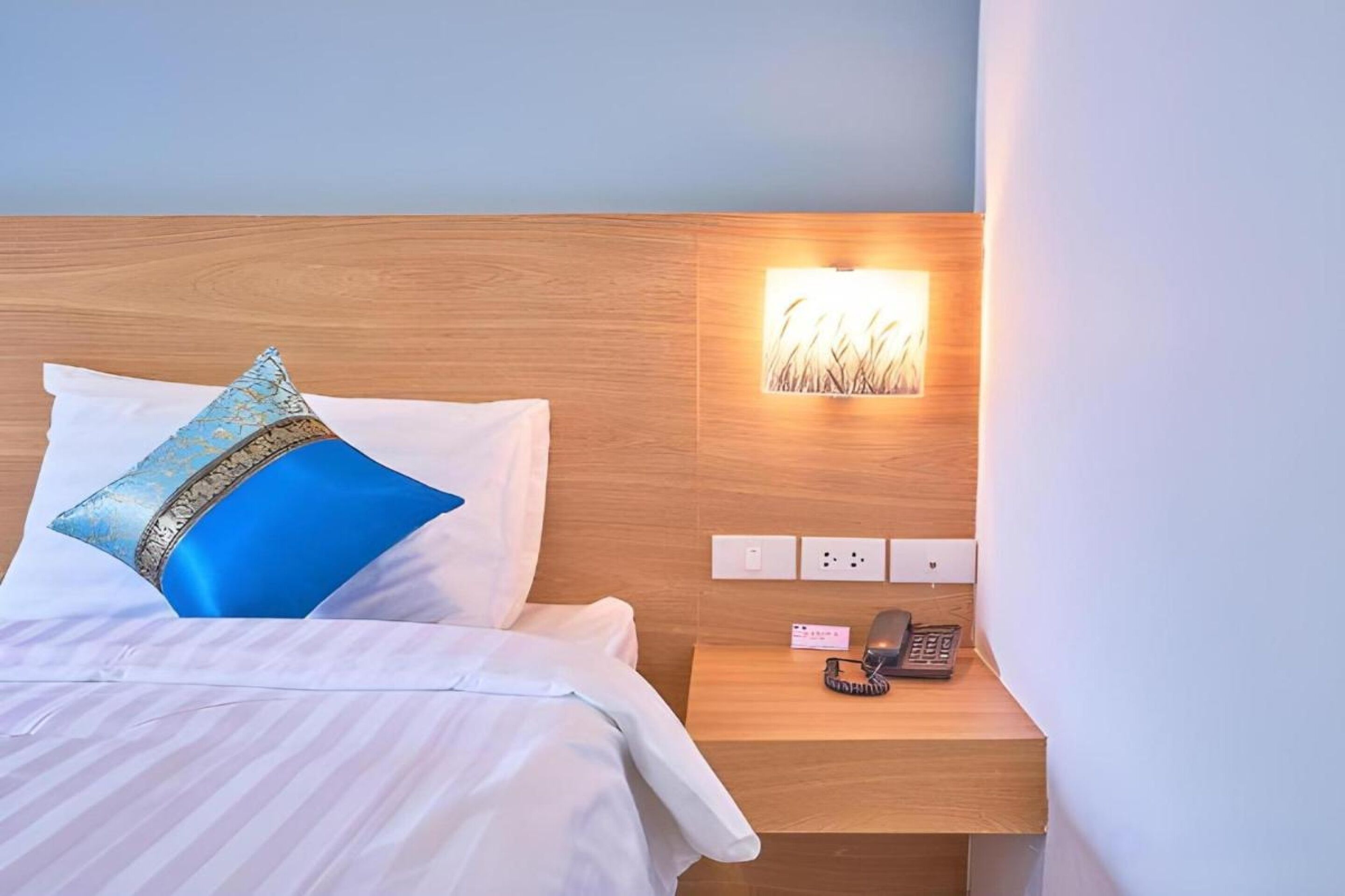 A hotel room with a neatly made bed, a bedside telephone, and a wall-mounted light fixture.