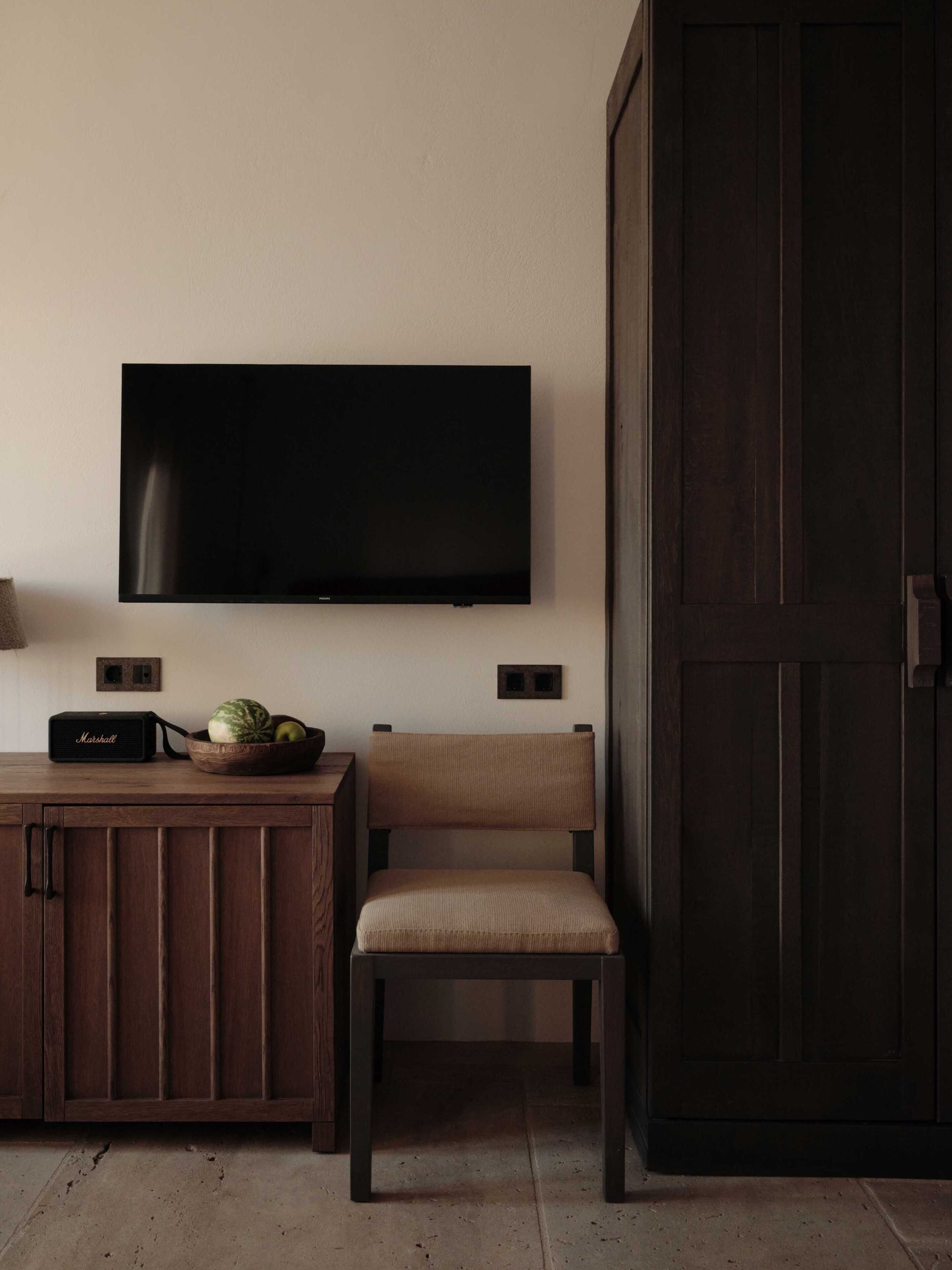 A room with a dark wooden door, a television mounted on the wall, a wooden cabinet with a Marshall speaker, and a bowl of fruit on the cabinet.