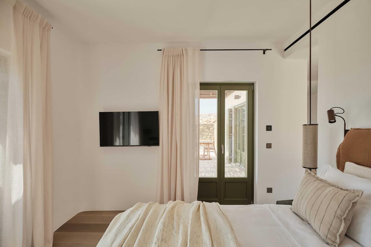A bedroom with a bed, a flat-screen TV mounted on the wall, a door leading to a balcony, and a lamp on the nightstand.