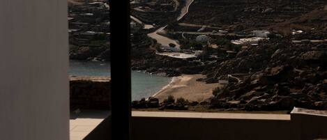 An open doorway leading to a terrace with a view of a coastal landscape, including a beach and a winding road.