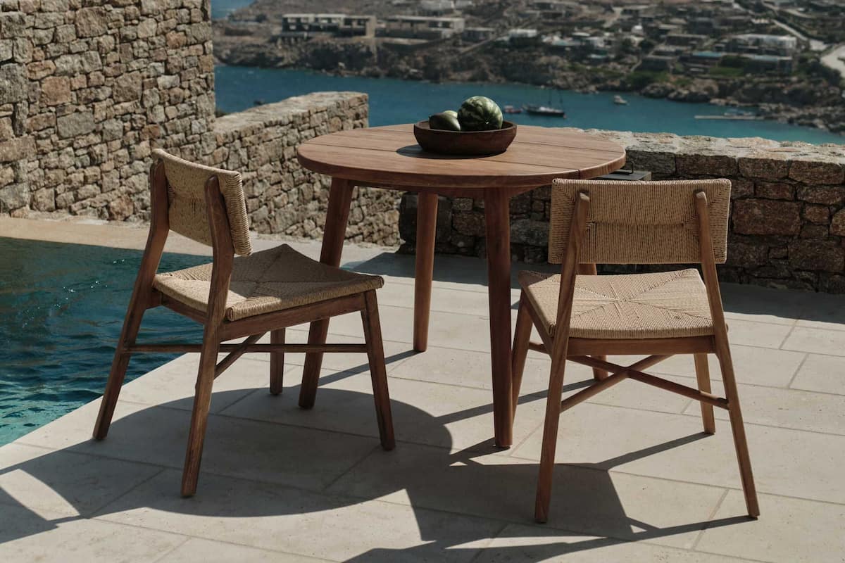 A patio with two wicker chairs and a round table, overlooking a coastal landscape with a stone wall and a pool.