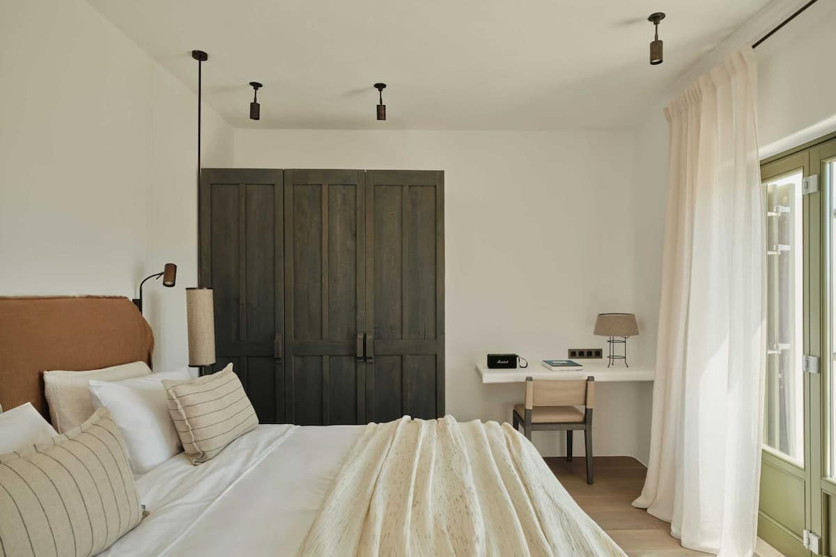 A bedroom with a bed, bedside lamps, a desk, a chair, and a large window with curtains.