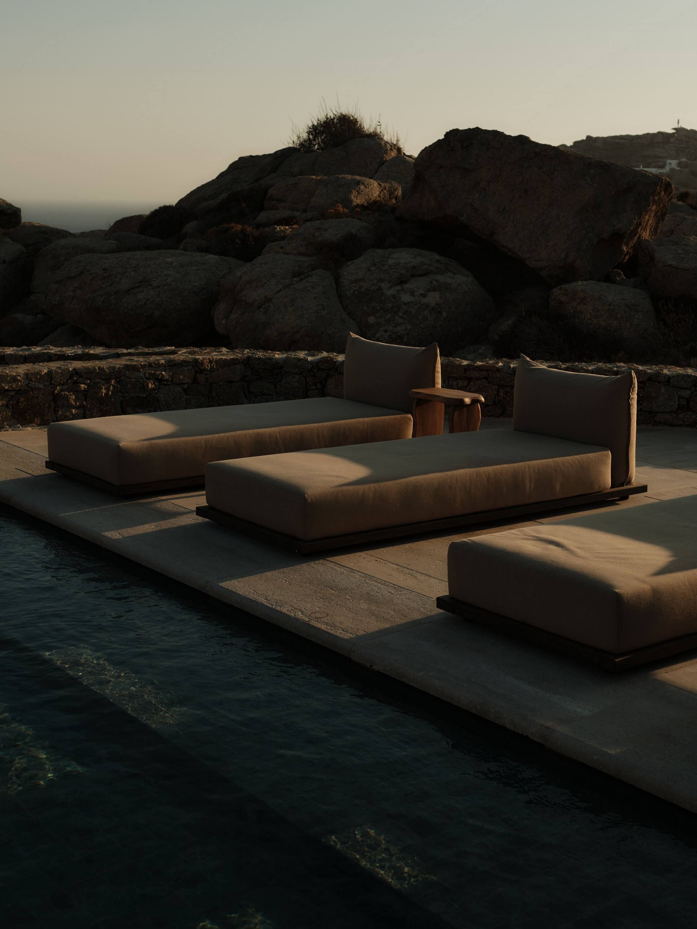 Zona junto a la piscina con tres espacios de descanso color beige y una pequeña mesa, frente a grandes rocas y un muro de piedra.