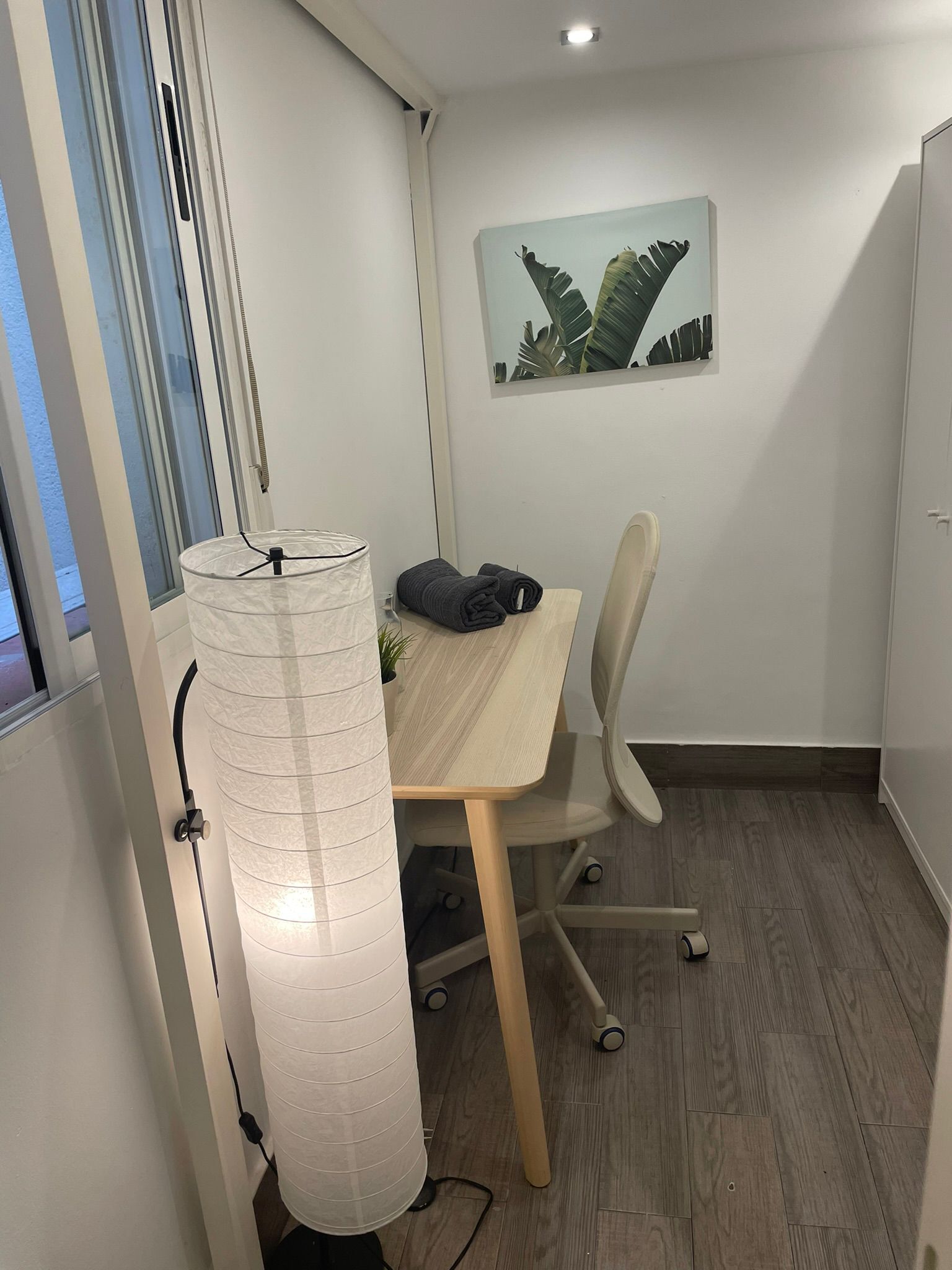 Een kleine kantoorruimte met een houten bureau, een witte stoel, een potplant en een aan de muur hangend schilderij.