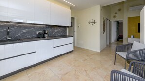Una cocina moderna con armarios blancos, encimera de mármol y cafetera. Hay un comedor con mesa y sillas.