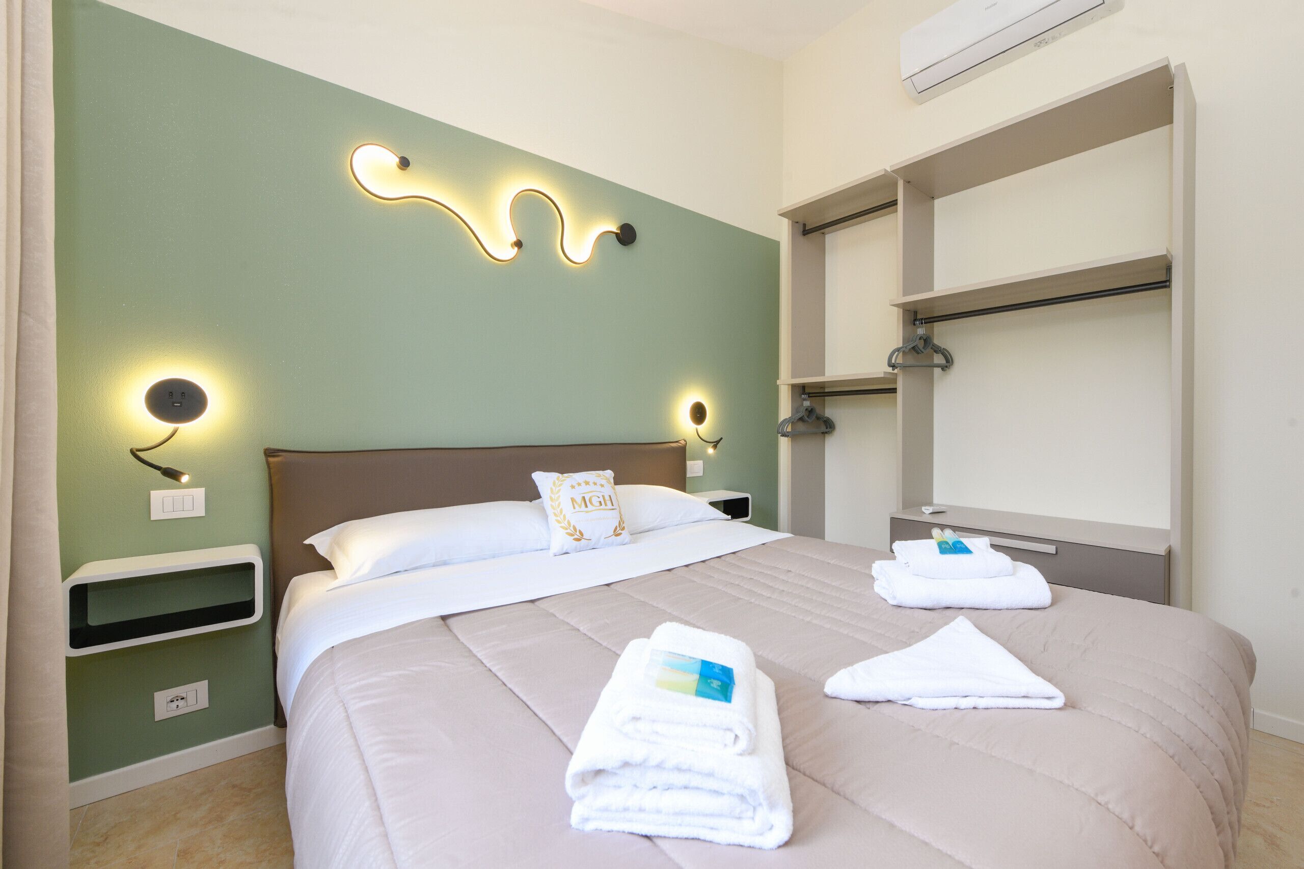 A neatly made bed with a beige comforter and pillows, a bedside table with a lamp, and a wall-mounted shelf with a towel.