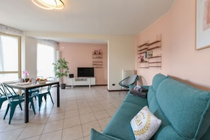 A living room with a teal sofa, a dining table with chairs, a TV, and a plant.