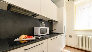 Eine Küche mit Mikrowelle, Schneidebrett mit Brot und Tomaten sowie einem Fenster mit Jalousien.