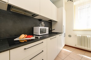 A kitchen with a microwave, a cutting board with bread and tomatoes, and a window with blinds.