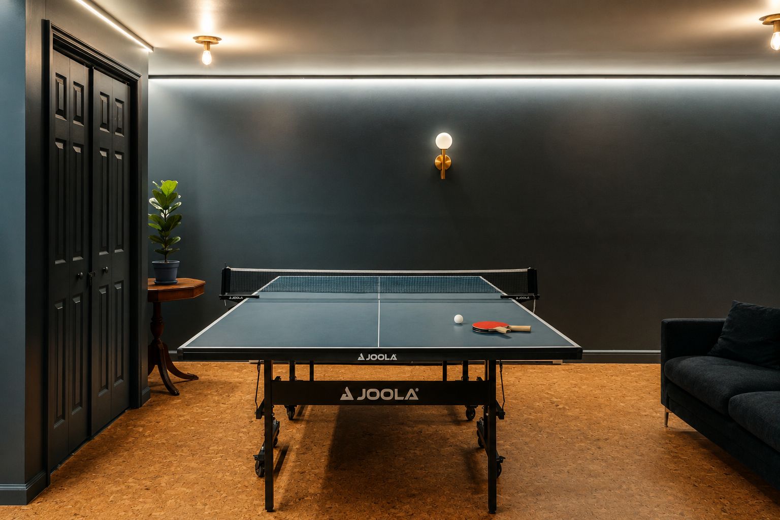 A ping pong table with a net, a paddle, and a ball in a room with dark walls, a potted plant, and a sofa.