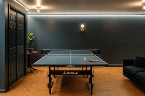 A ping pong table with a net, a paddle, and a ball in a room with dark walls, a potted plant, and a sofa. - Hot Tub, Fire Pit, Theater, Games & EV | Sleeps 8 (Barryville)