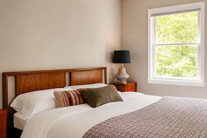 A bedroom with a wooden headboard, a bed with white bedding, a patterned throw blanket, a bedside lamp, and a window with a view of trees. - Hot Tub, Fire Pit, Theater, Games & EV | Sleeps 8 (Barryville)