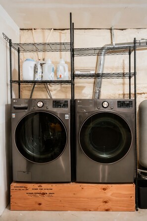 Two stacked front-loading washing machines with a metal shelf above them holding cleaning supplies. - Hot Tub, Fire Pit, Theater, Games & EV | Sleeps 8 (Barryville)