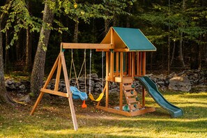 A wooden swing set with a slide, surrounded by trees and rocks. - Hot Tub, Fire Pit, Theater, Games & EV | Sleeps 8 (Barryville)