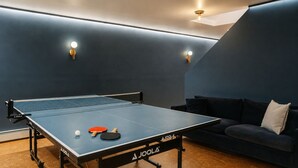 A ping pong table with a net, two paddles, and a ball in a room with a dark blue wall and a sofa.