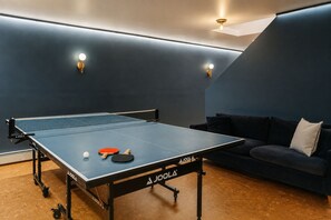 A ping pong table with a net, two paddles, and a ball in a room with a dark blue wall and a sofa.