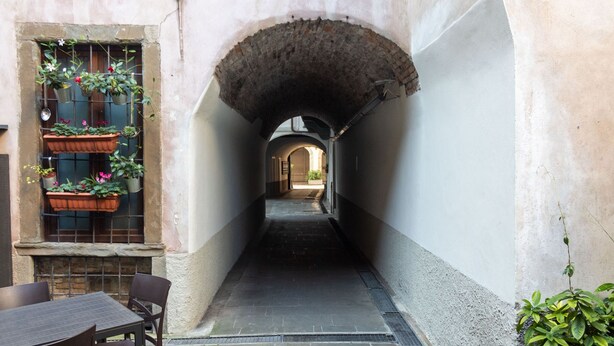 A narrow alleyway with a stone archway, potted plants on the left, and a table with chairs at the end.