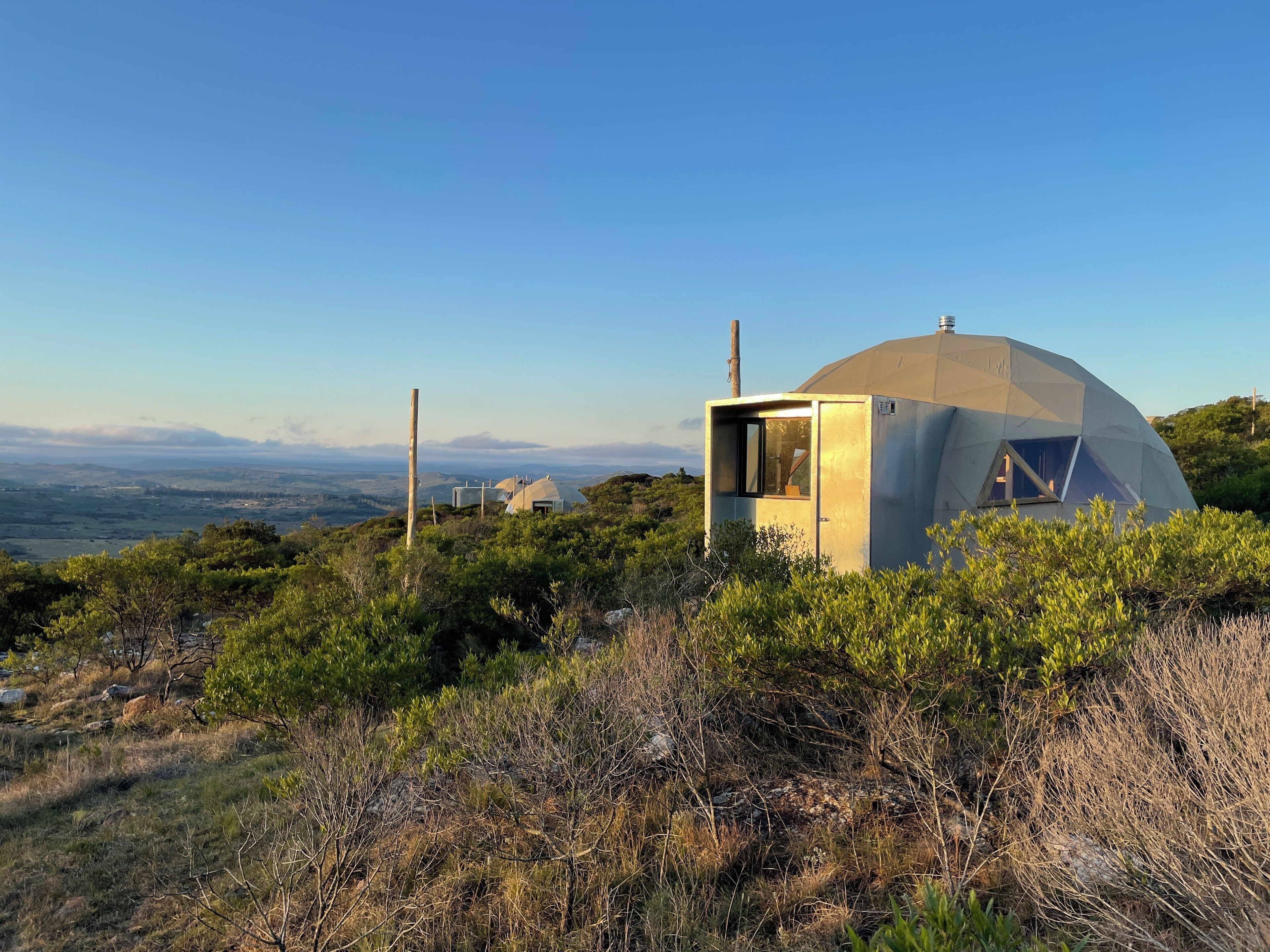 A geodesic dome structure with a small attached cabin, surrounded by dense shrubbery and hills in the background.