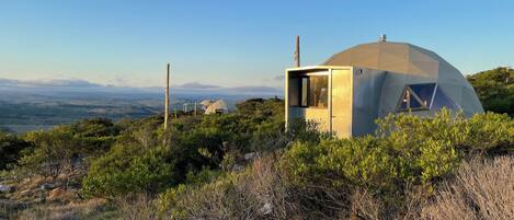 A geodesic dome structure with a small attached cabin, surrounded by dense shrubbery and hills in the background.