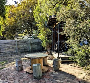 A rustic outdoor seating area with a round wooden table and stools, surrounded by trees and a wooden fence.
