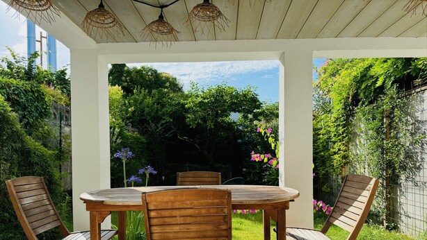 Une terrasse couverte avec une table et des chaises en bois, entourée d’une végétation luxuriante et offrant une vue sur des arbres et un bâtiment en arrière-plan.