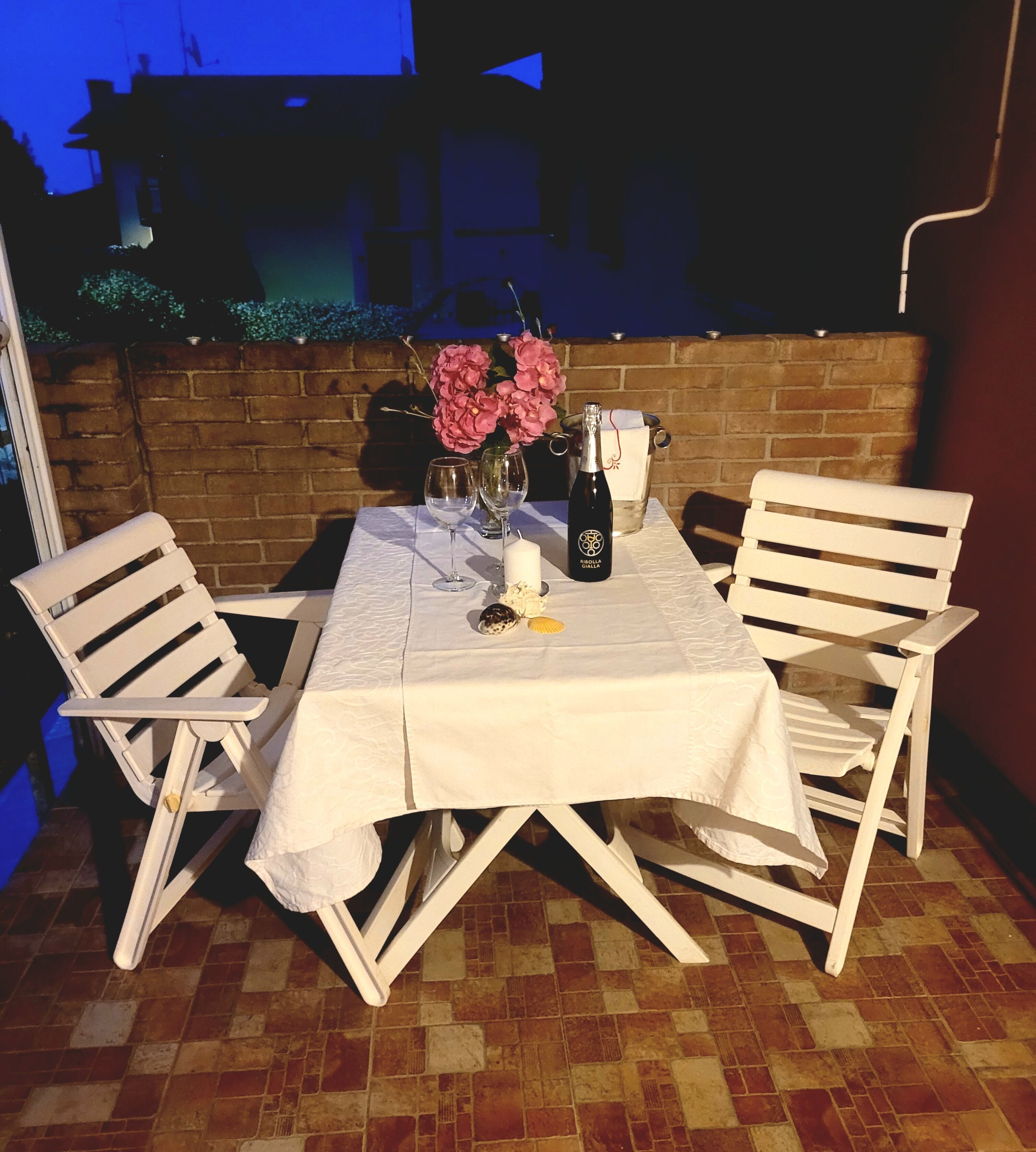 A table set outdoors with a white tablecloth, two chairs, a bottle of wine, a candle, and a vase with flowers.