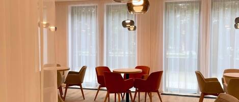 Un comedor moderno con mesas y sillas redondas, ventanas amplias con cortinas translúcidas e iluminación colgante.