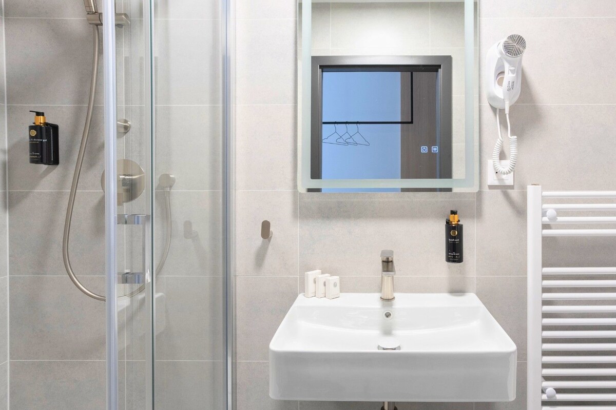 A modern bathroom with a glass shower enclosure, a white sink, a hairdryer mounted on the wall, and a towel rack.