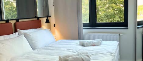 Ein ordentlich bezogenes Bett mit weißen Leinentüchern und zwei Handtüchern, ein Fenster mit Blick ins Grüne und eine Heizung an der Wand.