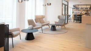 Een moderne woonkamer met een ronde salontafel, twee fauteuils en een bank. Er hangen hanglampen, een groot raam en in de achtergrond is een keuken te zien.